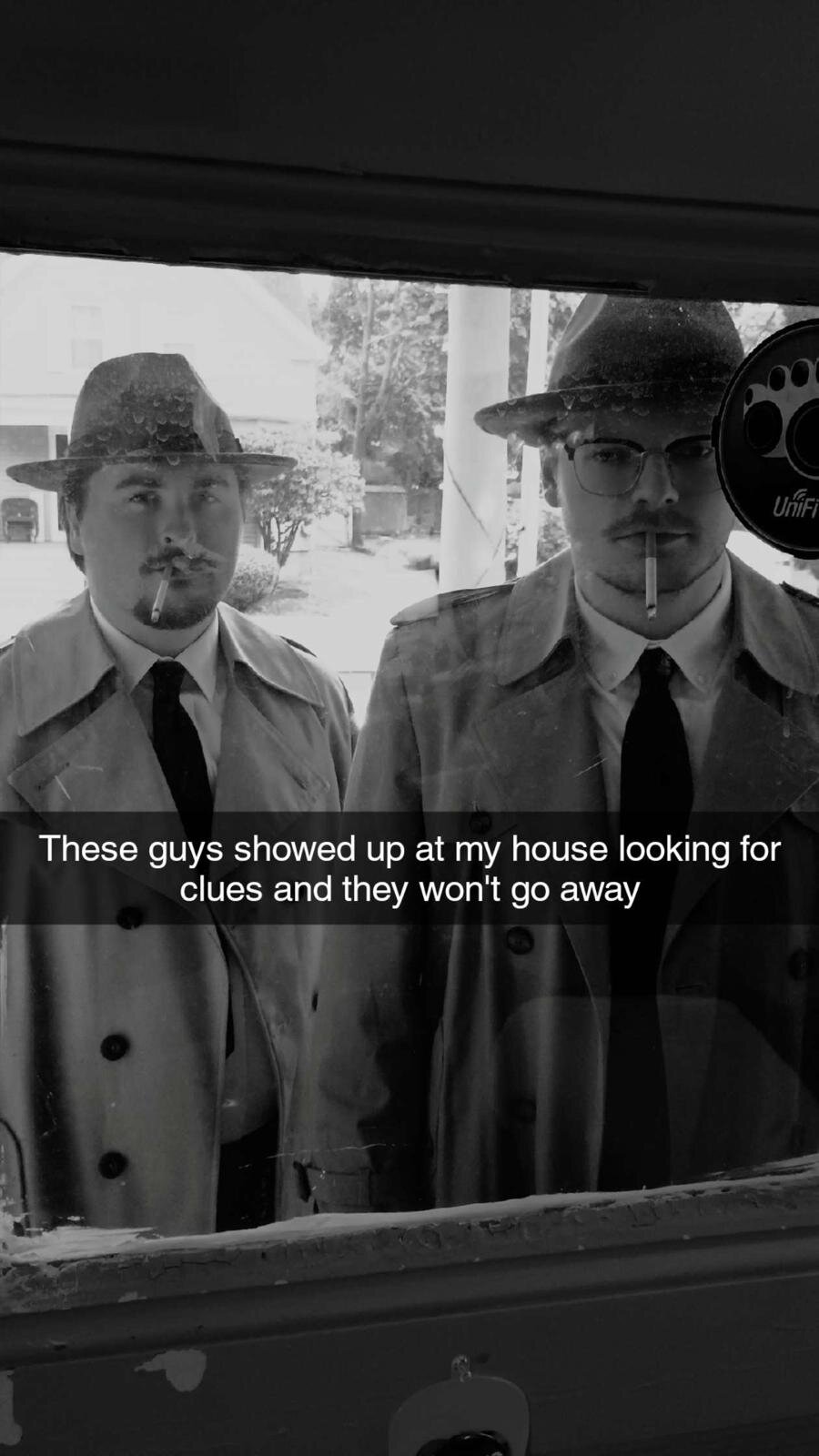Two detectives in trench coats and fedoras looking through a window